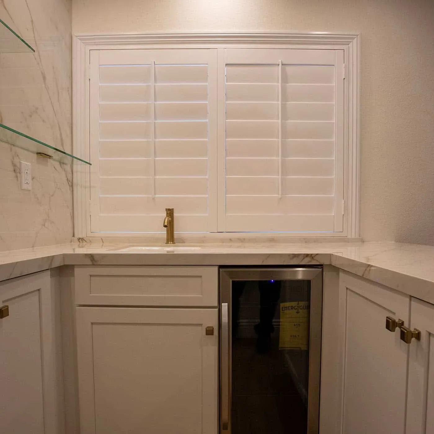 Home bar with white shaker cabinets, a veined white counter, a brass faucet, a wine fridge and glass shelves