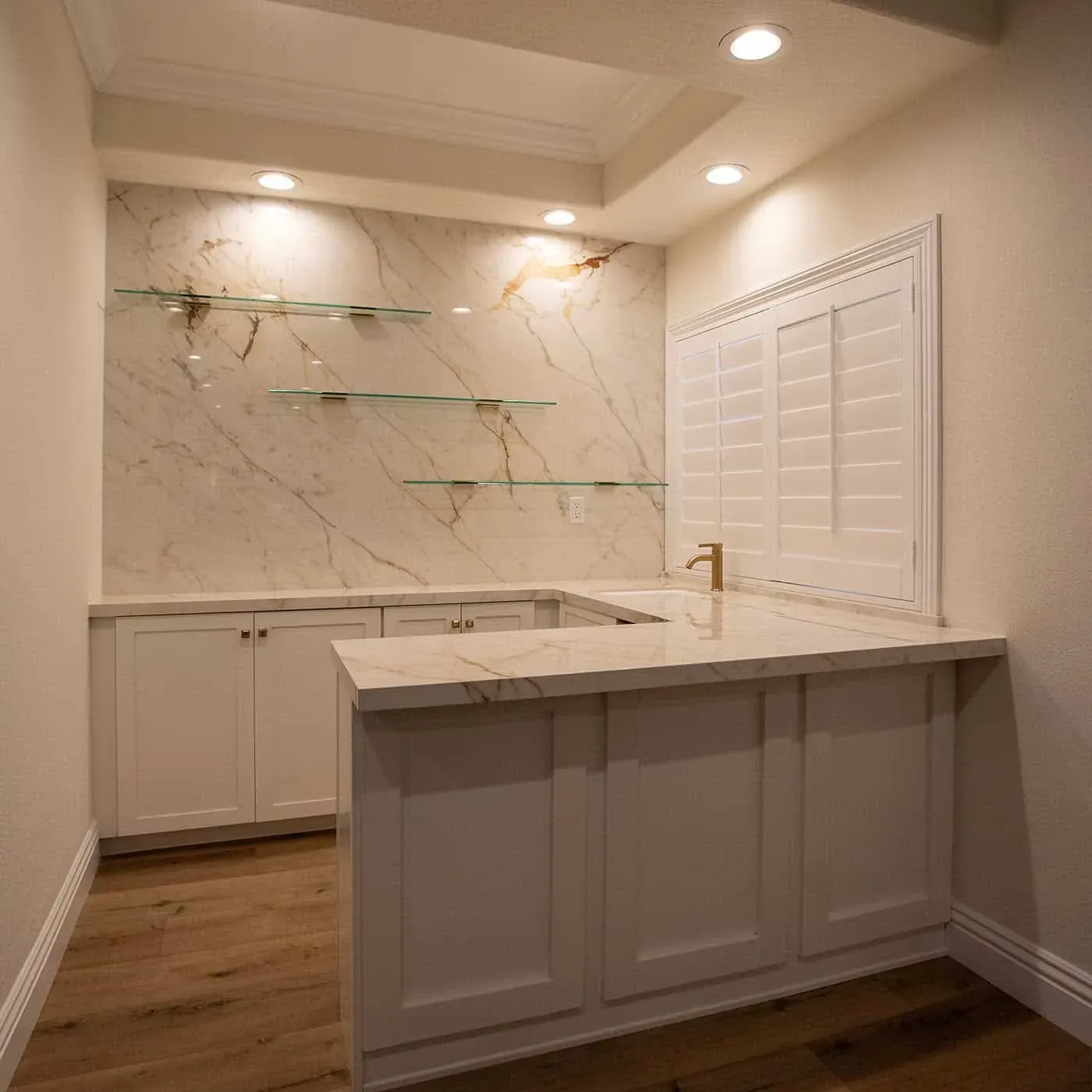 Bar counter with floating glass shelves set against a book-matched marble slab wall and a brass tap