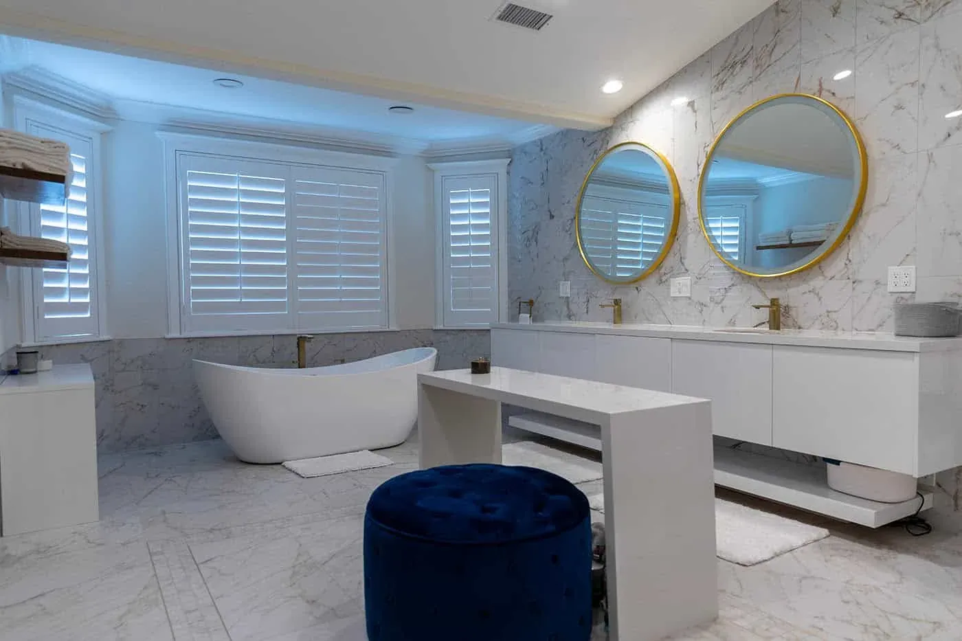 Marble bathroom with round gold mirrors, plantation shutters and a freestanding tub