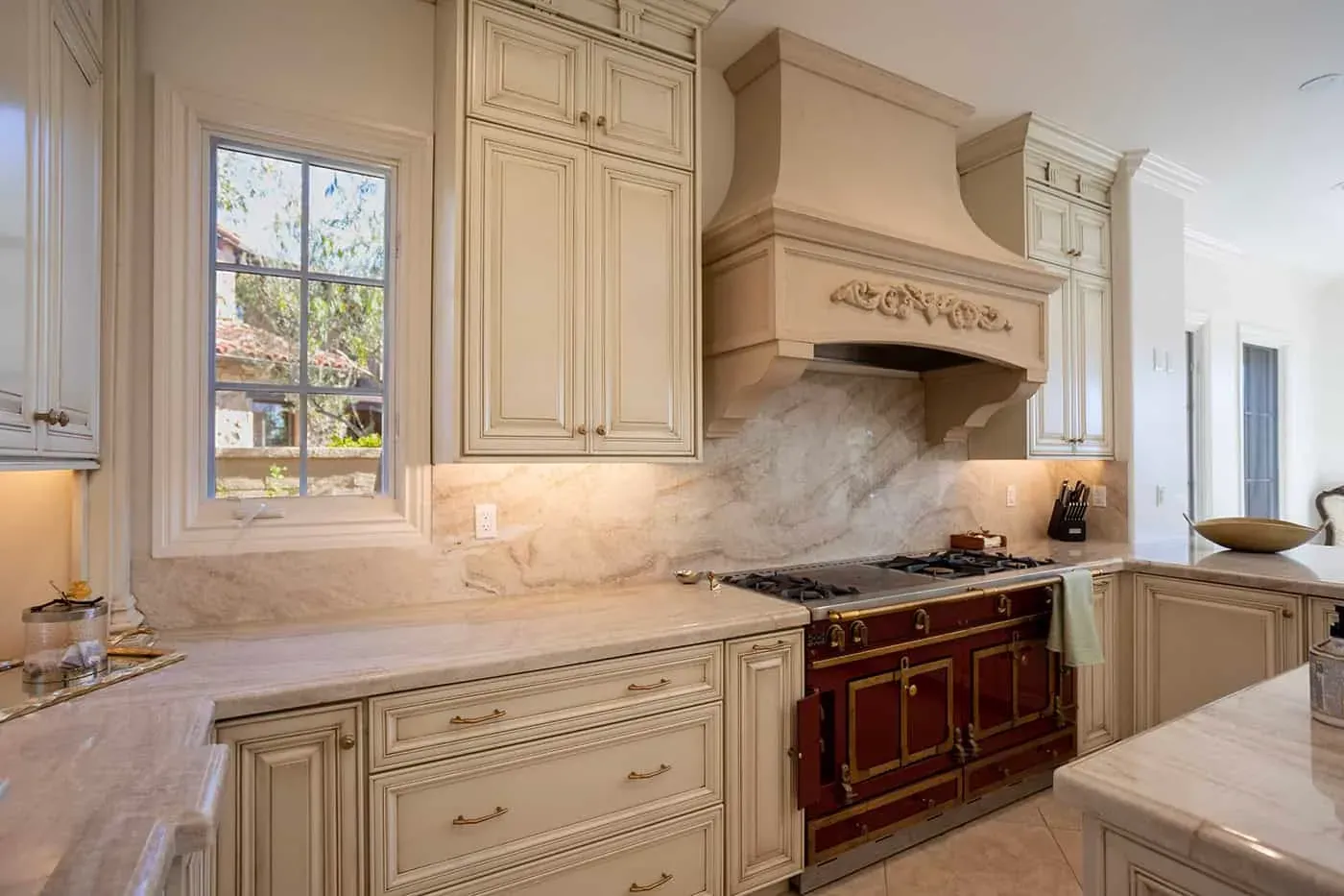 Newport Beach kitchen with a carved hood above a red range beside a window and stone backsplash