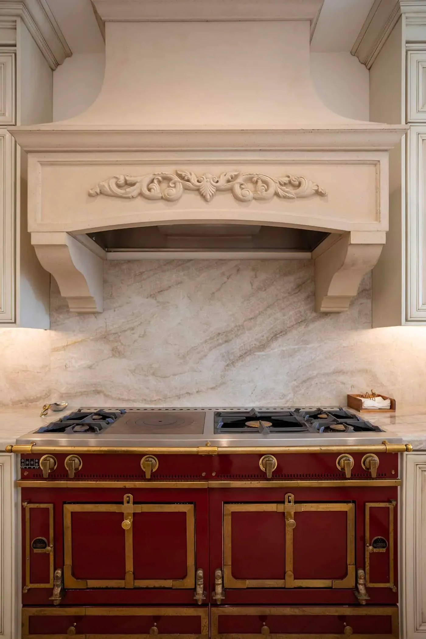 Carved hood above a red range with a book-matched stone backsplash