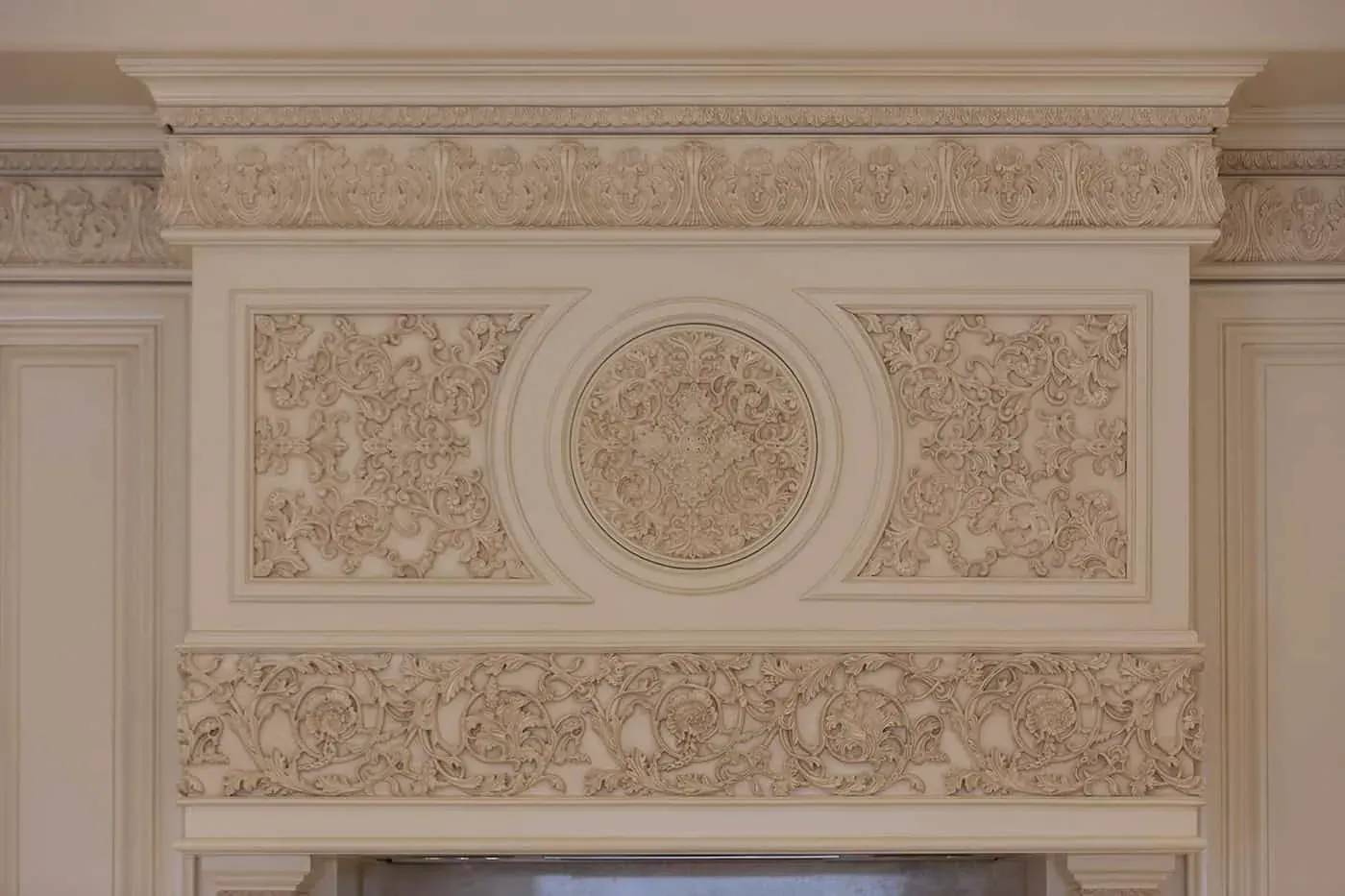 Cream painted range hood overmantel with carved scrollwork panels, a central carved medallion and a leaf-carved crown moulding