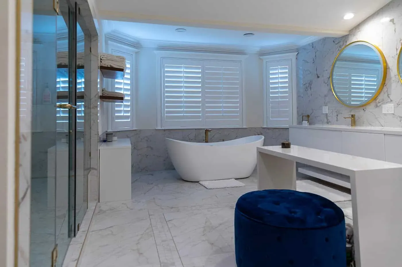 Marble bathroom with a glass shower, a freestanding tub under plantation shutters and round gold-framed mirrors