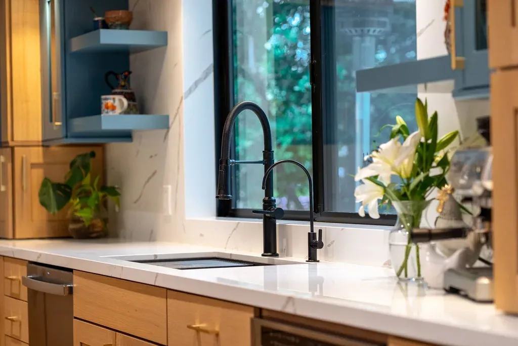 Matte black pull-down faucet over an undermount sink in white quartz below a black-framed window