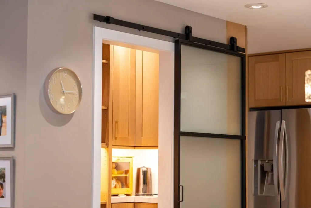 Sliding barn door with frosted glass panels in a black steel frame, open onto a lit oak pantry
