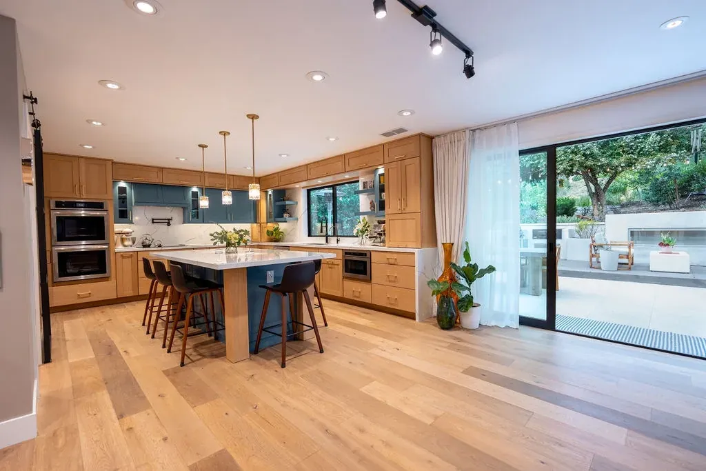Open kitchen with natural oak shaker cabinets, a teal island base under a white quartz top and sliding doors to the garden