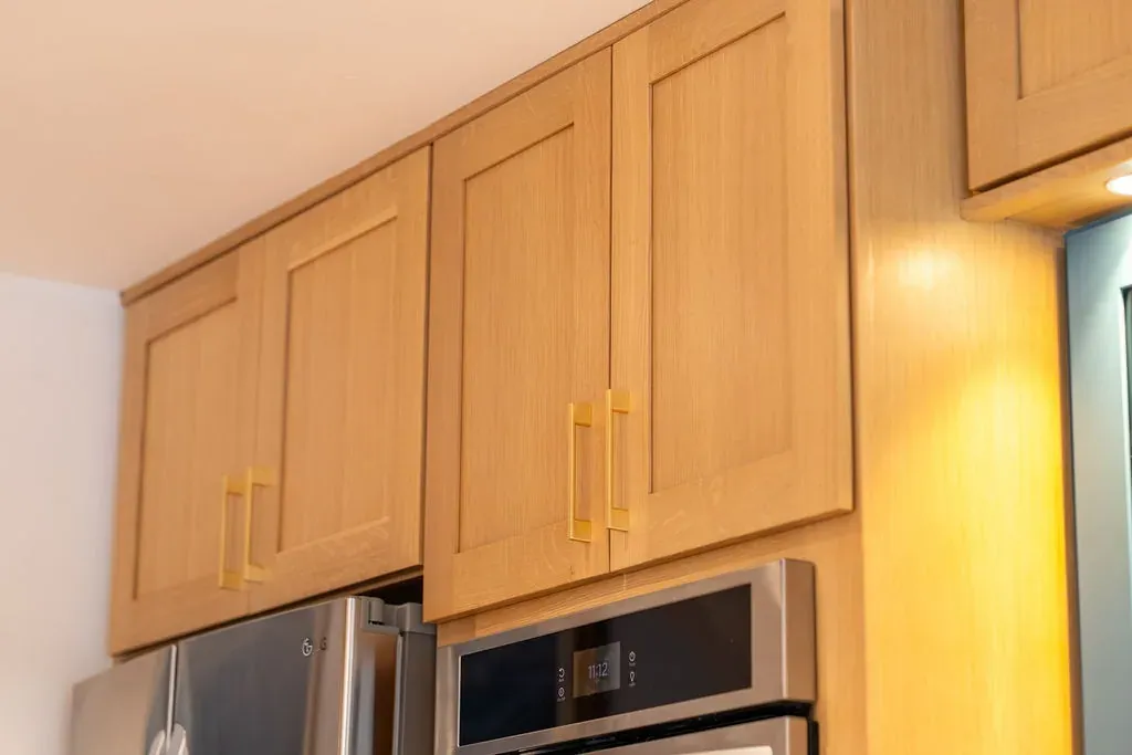 Natural oak shaker wall cabinets with long brass pulls, warmly lit, above a stainless microwave