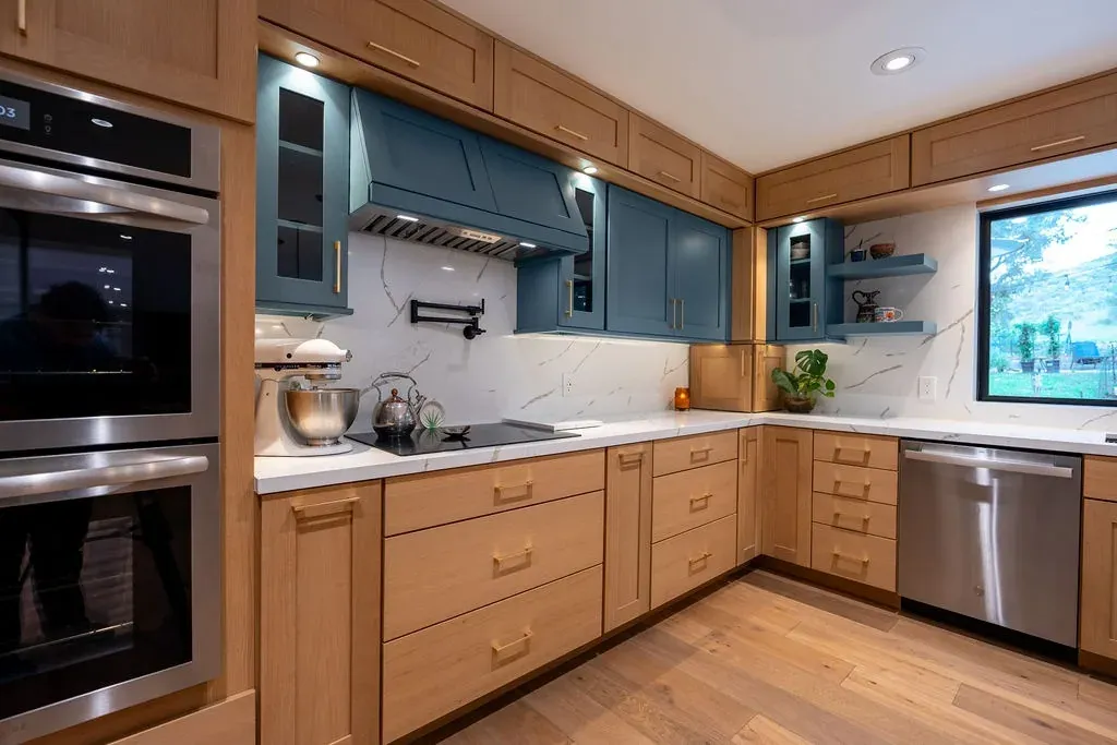 Teal painted range hood and wall cabinets over a cooktop, beside stainless double wall ovens and a veined white quartz backsplash