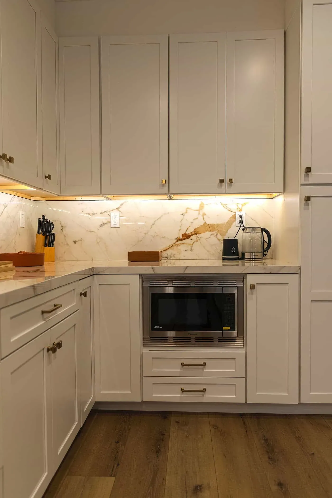 Cream shaker cabinetry with a built-in microwave under a marble backsplash lit from beneath the uppers