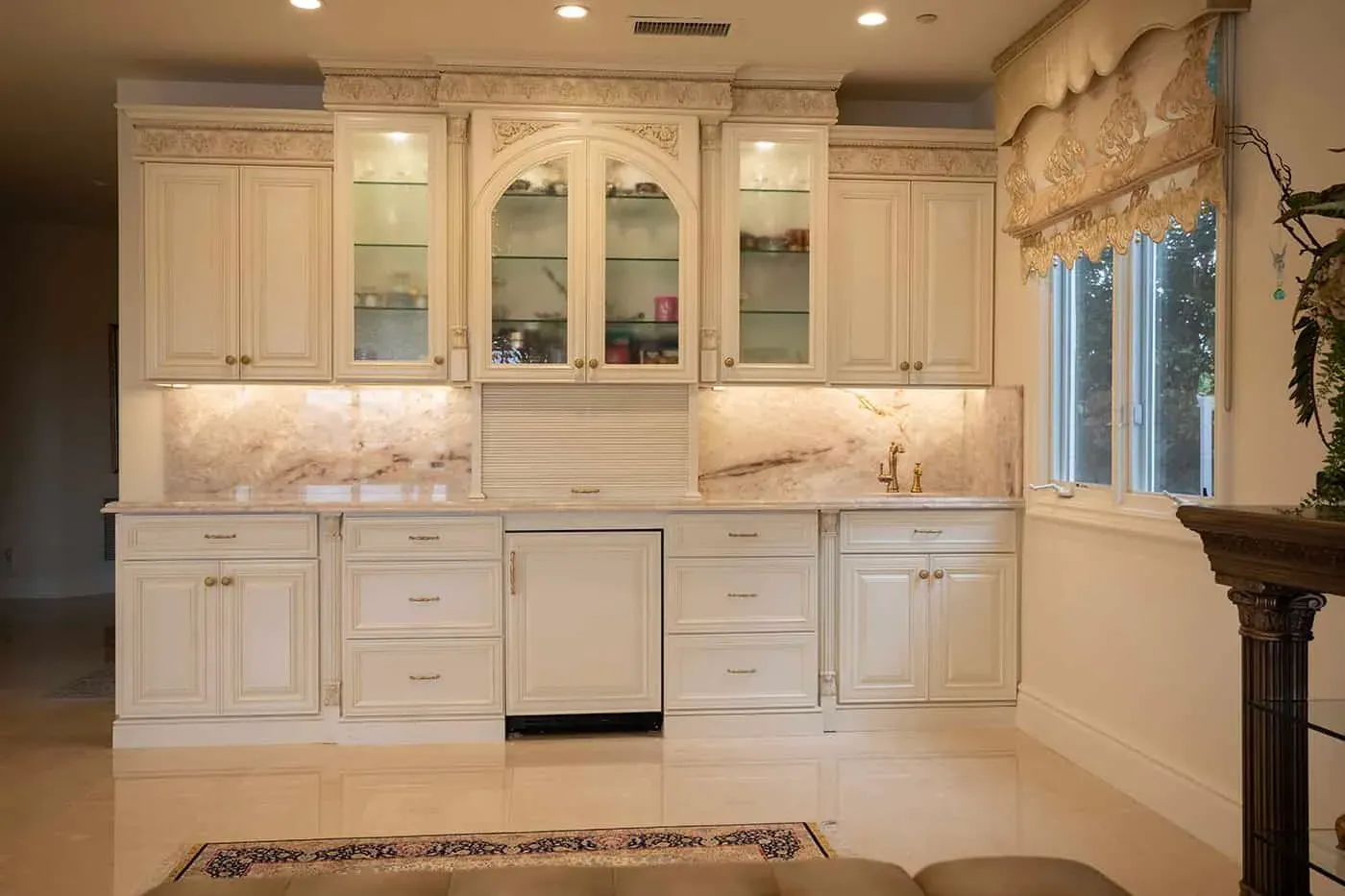 Cream cabinetry with glass-front uppers, a panelled dishwasher and an onyx backsplash lit from under the cabinets