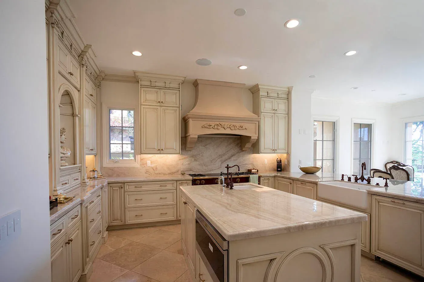 Cream traditional kitchen with a carved plaster range hood, a veined stone island and a farmhouse sink beneath the window