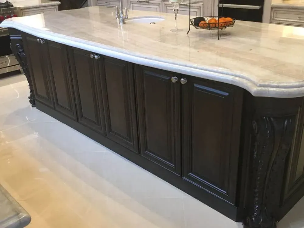 Dark carved kitchen island with a thick pale stone top and a rounded ogee edge on a polished floor