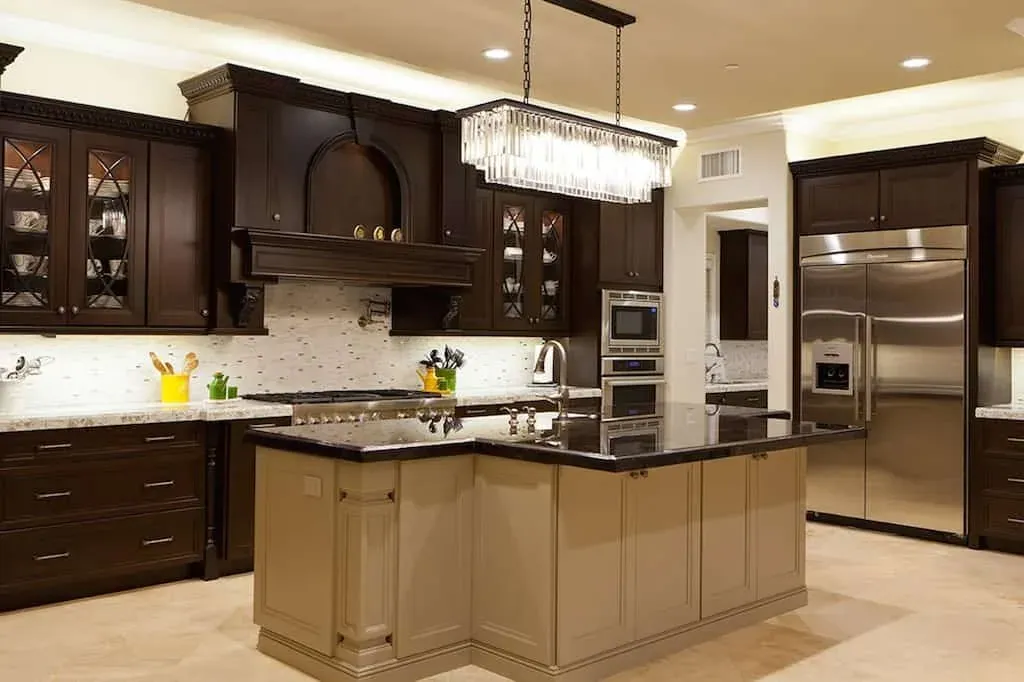 Dark kitchen with a linear chandelier over a granite island and wall ovens