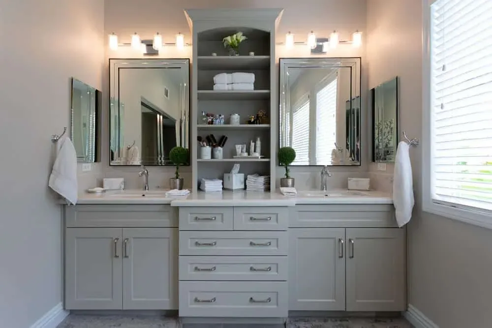 Grey double vanity with two framed mirrors, wall sconces and an open shelving tower between the sinks
