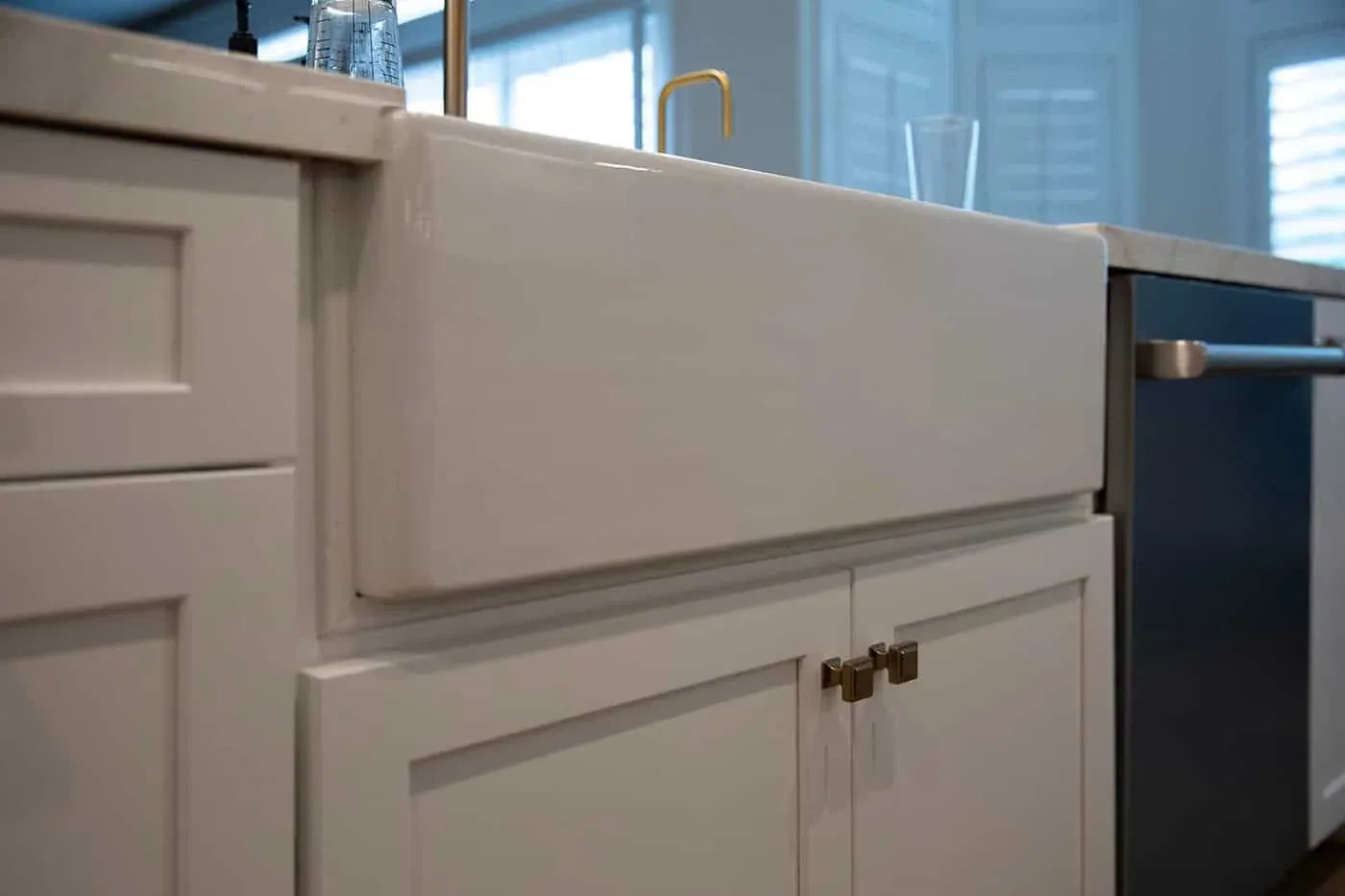 Farmhouse apron sink set into white shaker cabinetry with brass knobs and a brass tap