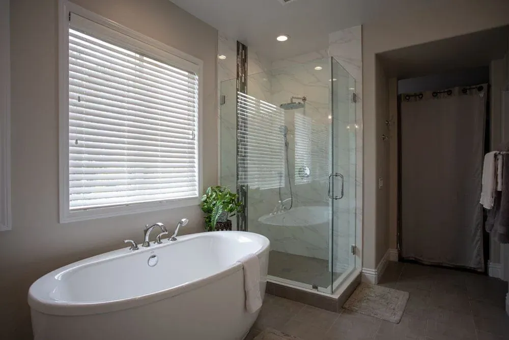 Oval freestanding tub under shuttered windows beside a corner glass shower with a mosaic accent strip