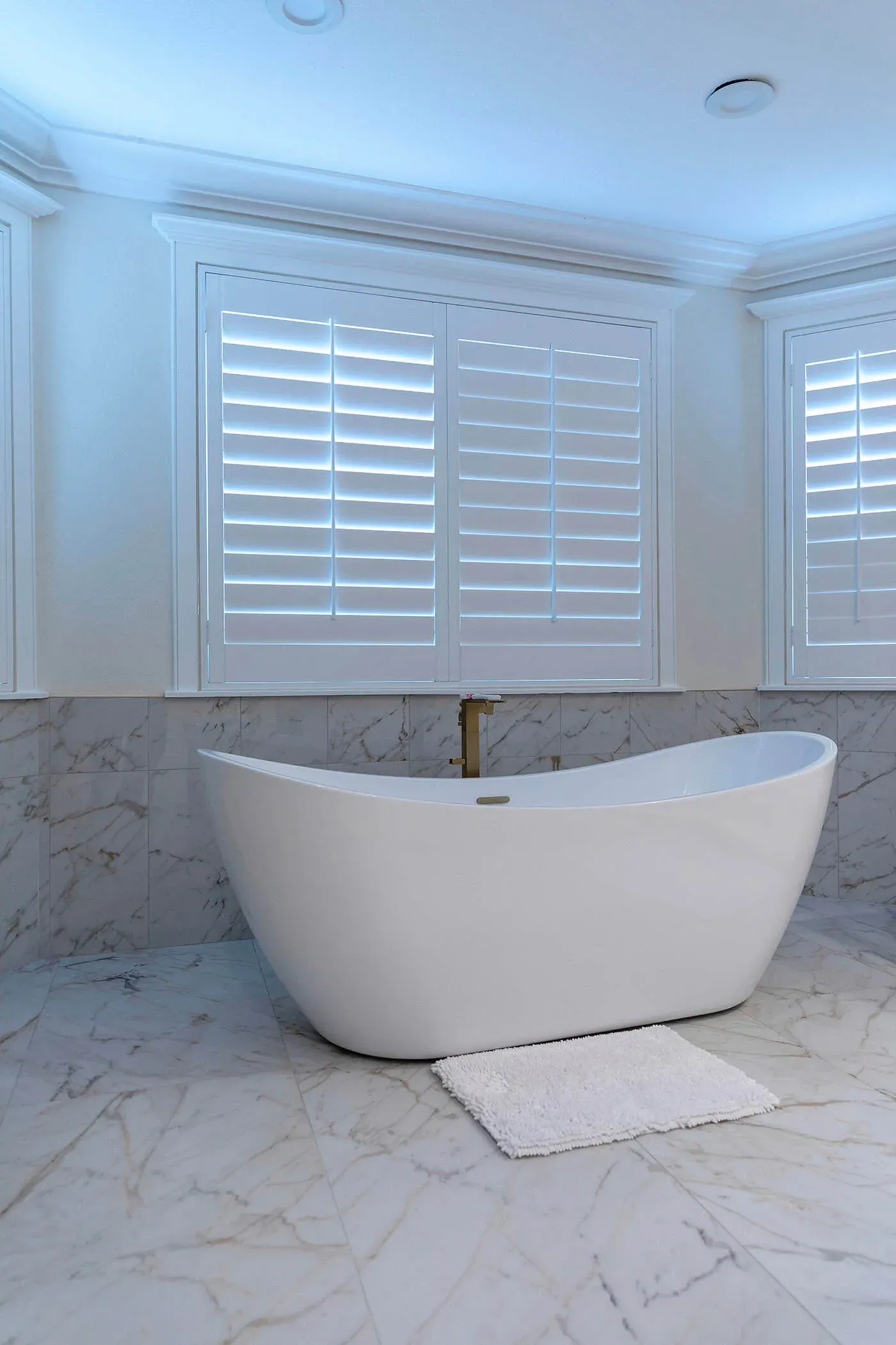 Freestanding tub beneath plantation shutters against a marble wall