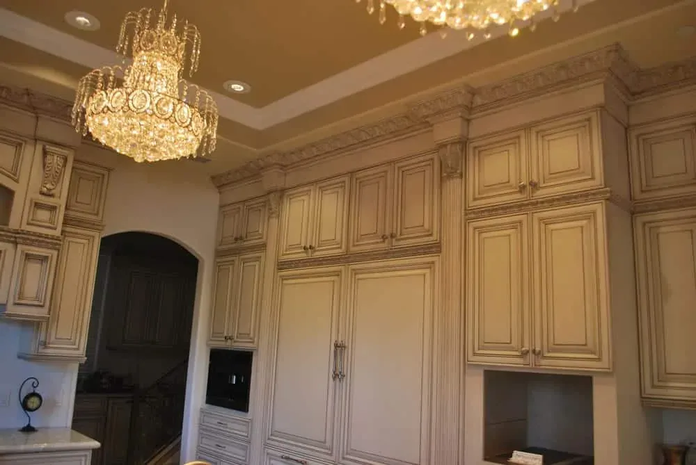 Floor-to-ceiling glazed cream cabinetry with carved crown moulding beside an arched doorway under crystal chandeliers