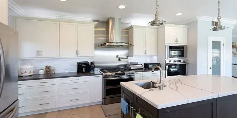 White kitchen with stainless range hood, dark quartz waterfall island and light countertops throughout