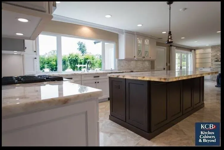 Dark wood kitchen island with waterfall marble countertop, white perimeter cabinets and a bay window above the sink