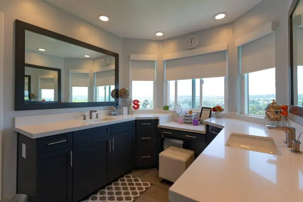 Bathroom vanity run with navy cabinets, a white quartz top and a wide mirror under a bay window