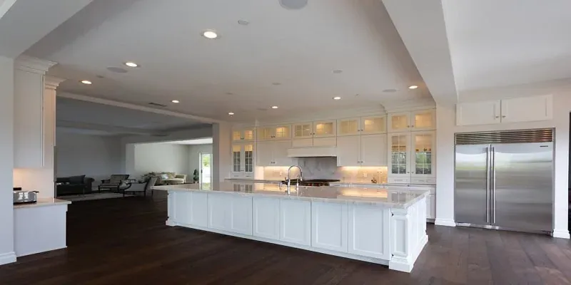 Long white kitchen island with gray countertops, dark hardwood flooring and a stainless side-by-side refrigerator