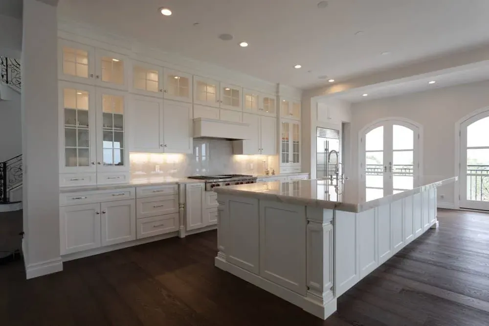 White kitchen with glass-front upper cabinets, a long white island and dark wood floors