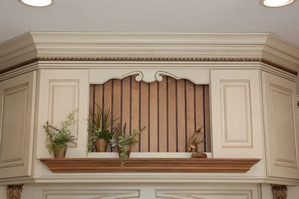 Carved overmantel shelf above cream cabinetry with potted plants
