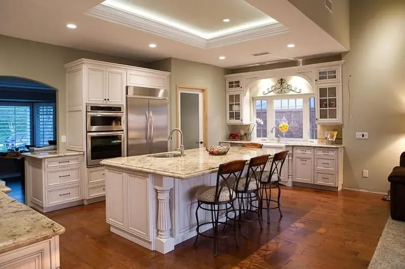 White kitchen with a granite-topped island, bar stools, stainless refrigerator and wood flooring under a tray ceiling