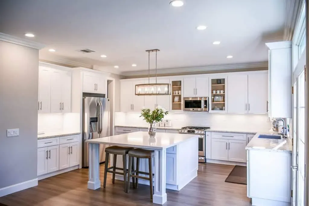 White shaker kitchen with a long white island, linear pendant light, stainless refrigerator and wide-plank wood floor