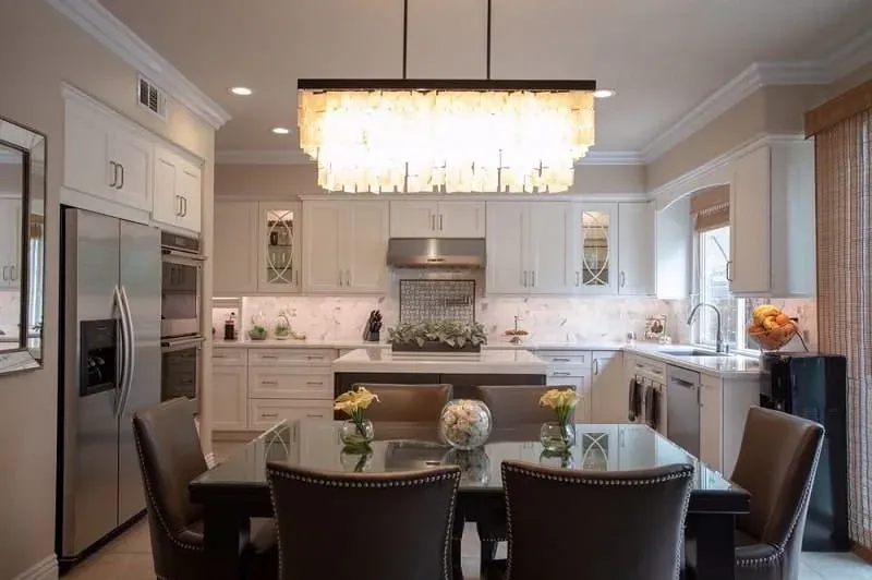 white kitchen with stainless steel refrigerator and wall ovens beneath a large rectangular chandelier over a glass dining table