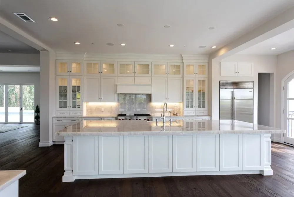Long white kitchen island with marble-look countertop, glass-front upper cabinets and dark wood flooring