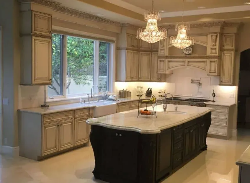 Antiqued cream cabinets with a farmhouse sink, two crystal chandeliers and a dark wood island topped with white marble
