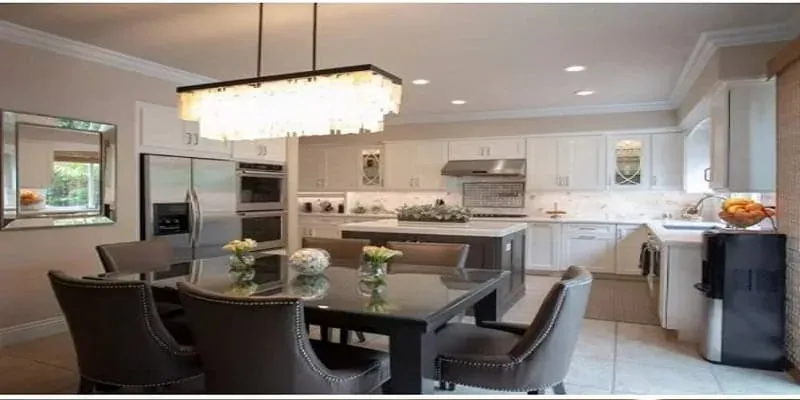 Dark upholstered dining table set beneath a crystal chandelier near a white kitchen with an island