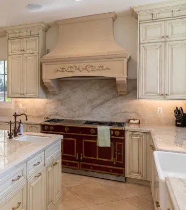 Cream kitchen with an ornate carved wood range hood, marble backsplash and a burgundy range with brass trim