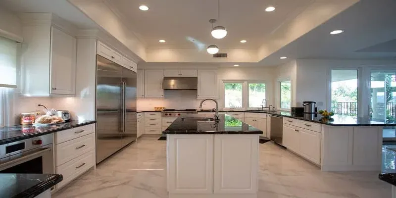 Bright white kitchen with a large island, black countertops, a stainless refrigerator and marble-look floor tile