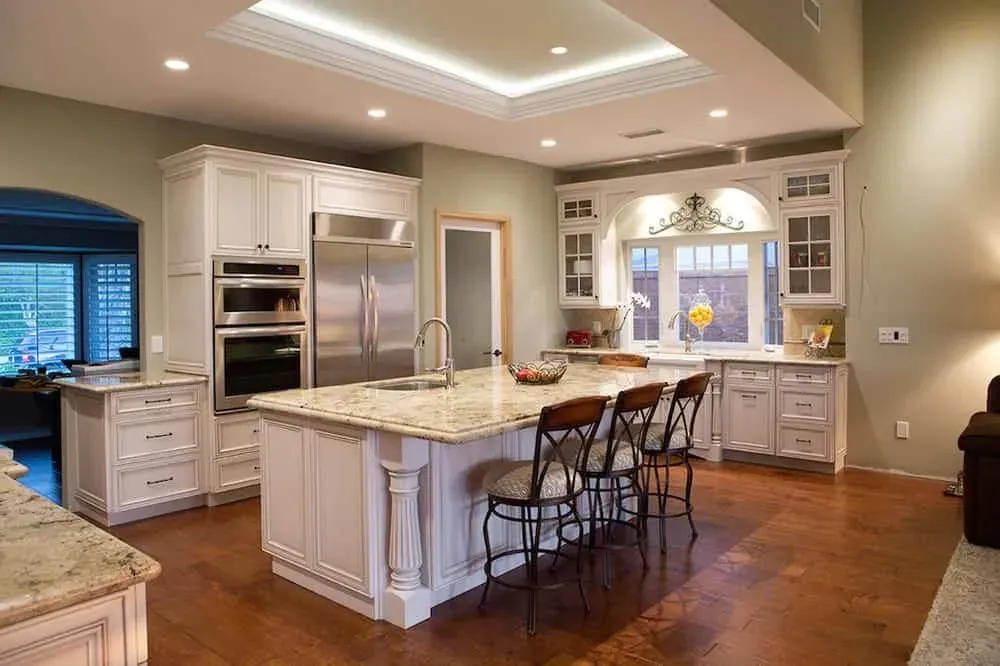 White kitchen with a granite island, bar stools and a lit tray ceiling over wood floors