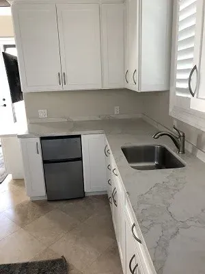 White kitchen corner with a small stainless mini fridge, light stone-look counters and plantation shutter window