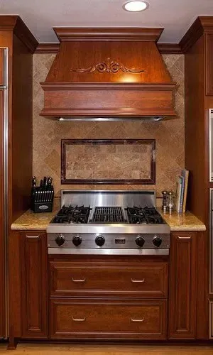 Carved wood range hood above a stainless cooktop with a framed tile backsplash and granite counter