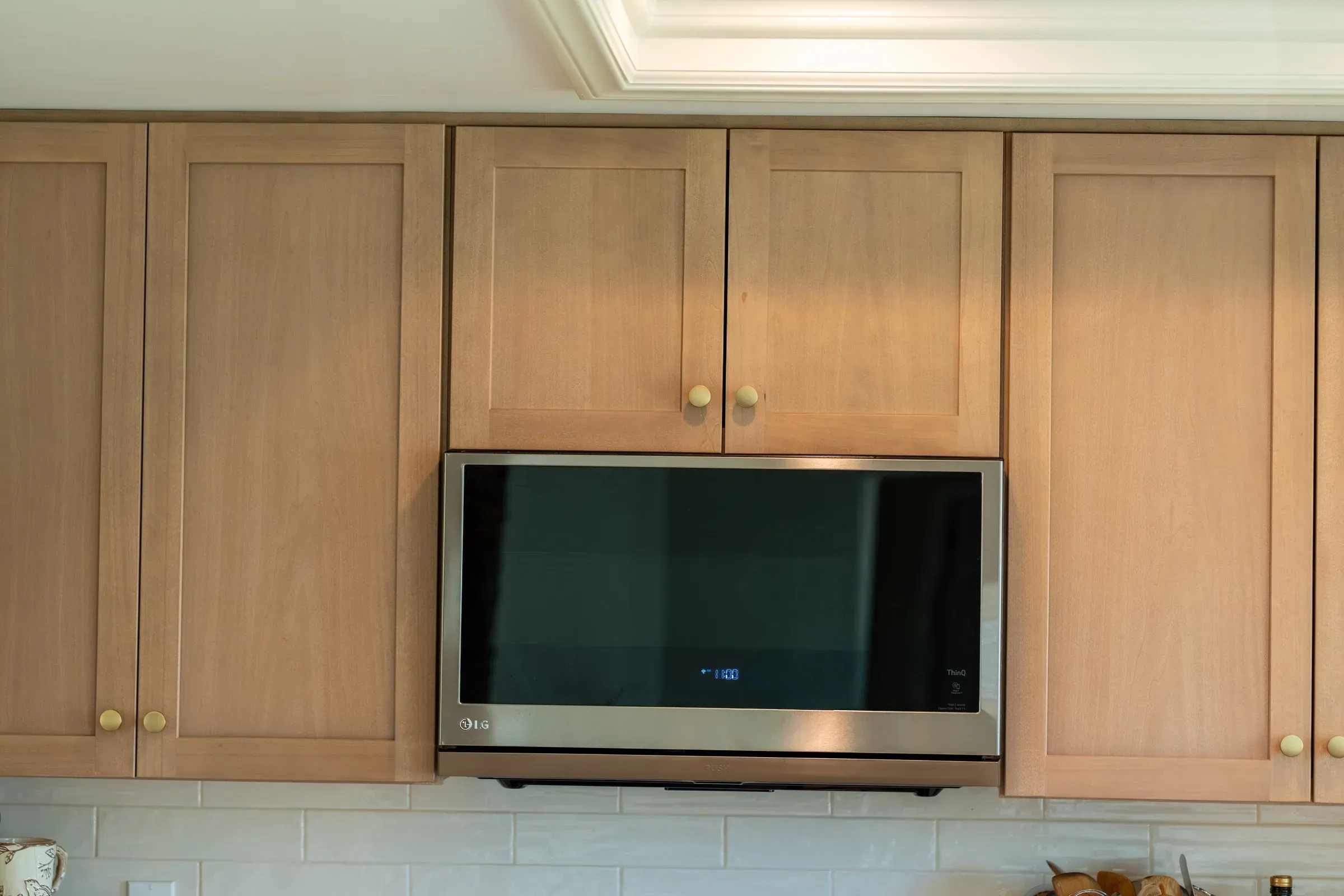 Light oak shaker wall cabinets with brass knobs framing a built-in stainless microwave above white tile