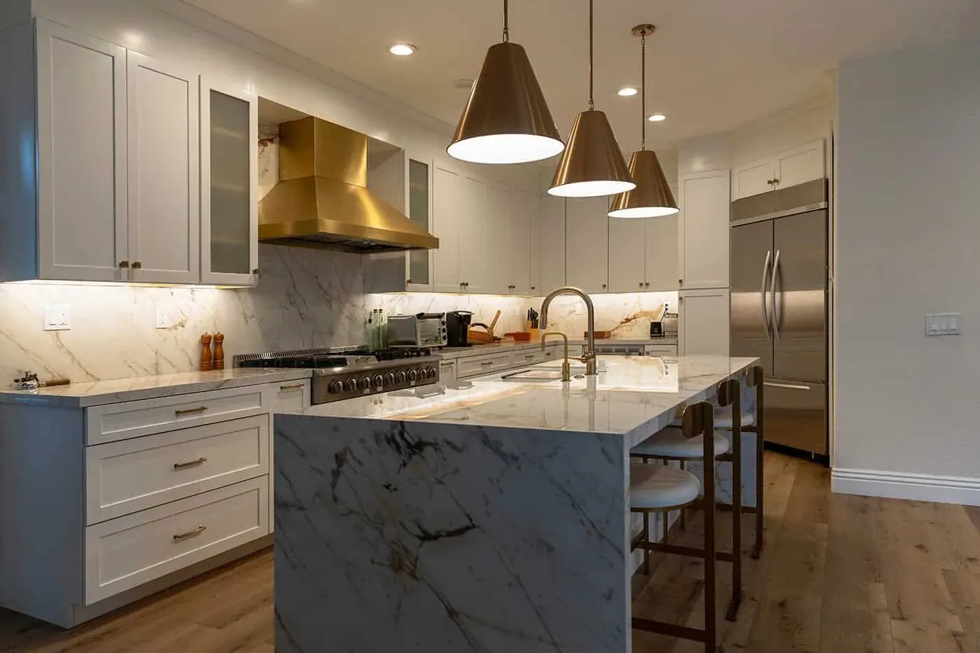 Waterfall marble island under three brass pendants beside a stainless refrigerator and a brass range hood