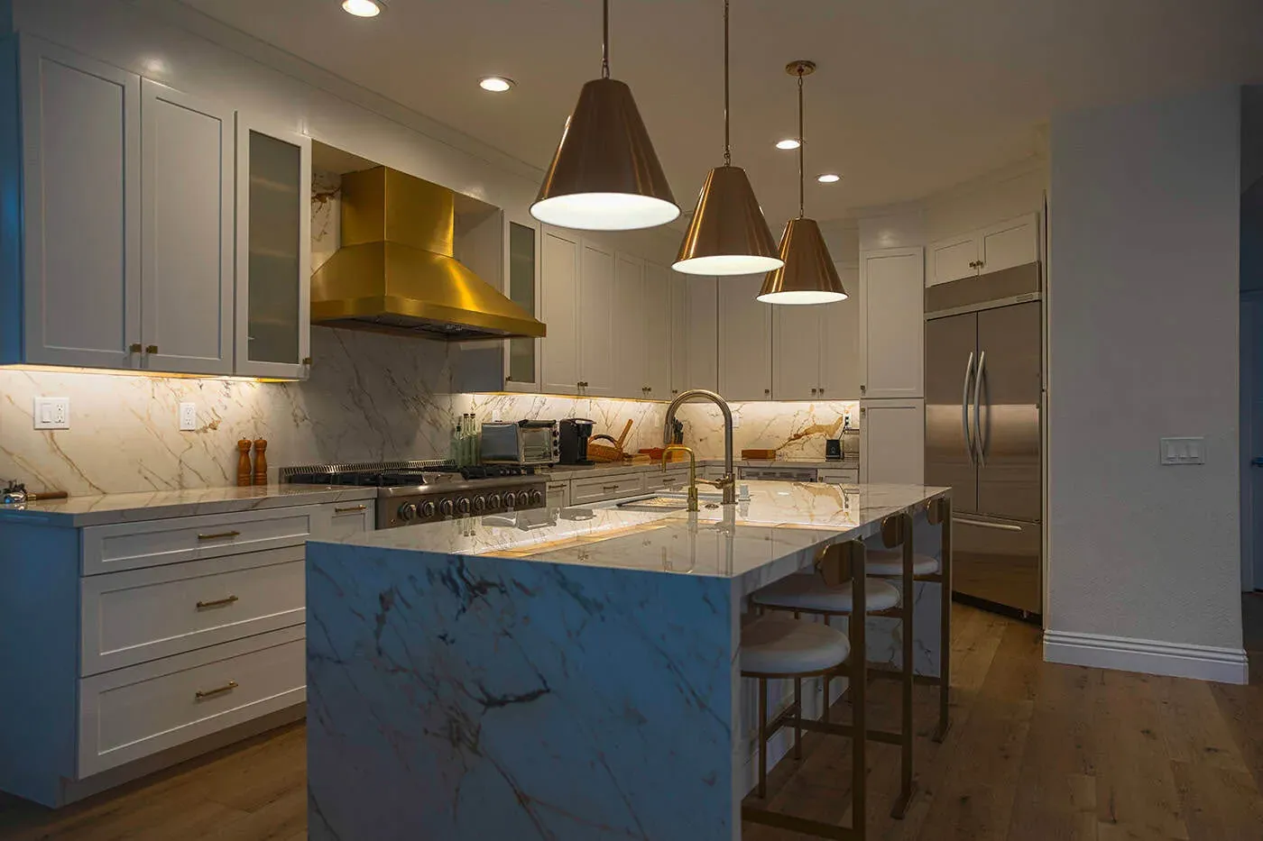 Wood plank floor meeting a blue marble island under three brass pendants in a white kitchen