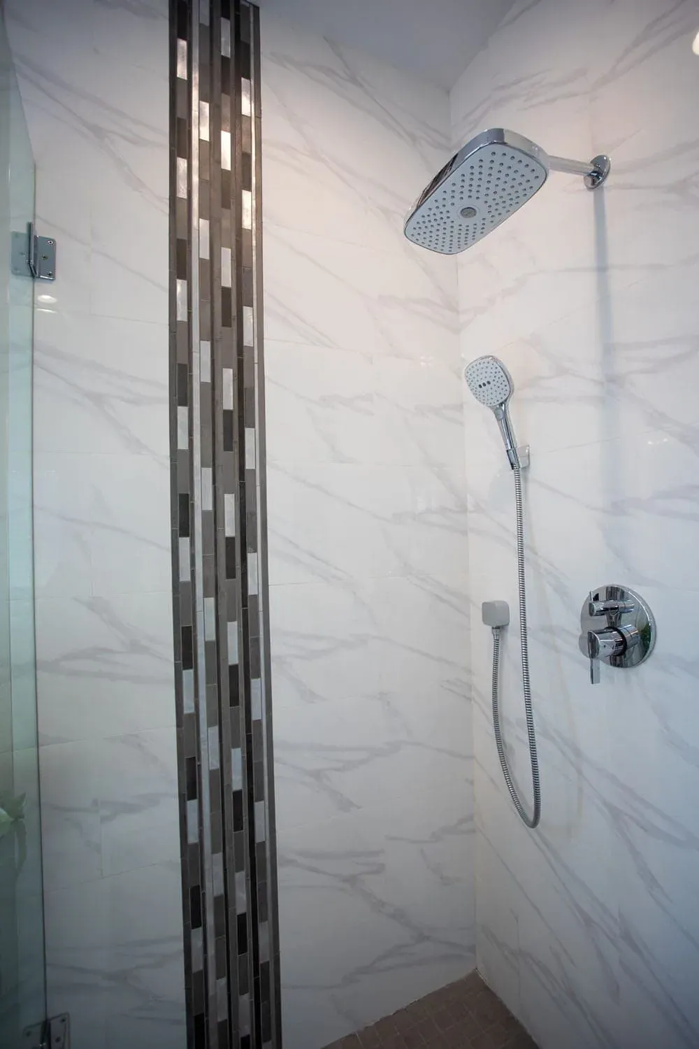 Marble-look tiled shower wall with a vertical mosaic accent strip and a chrome handheld shower