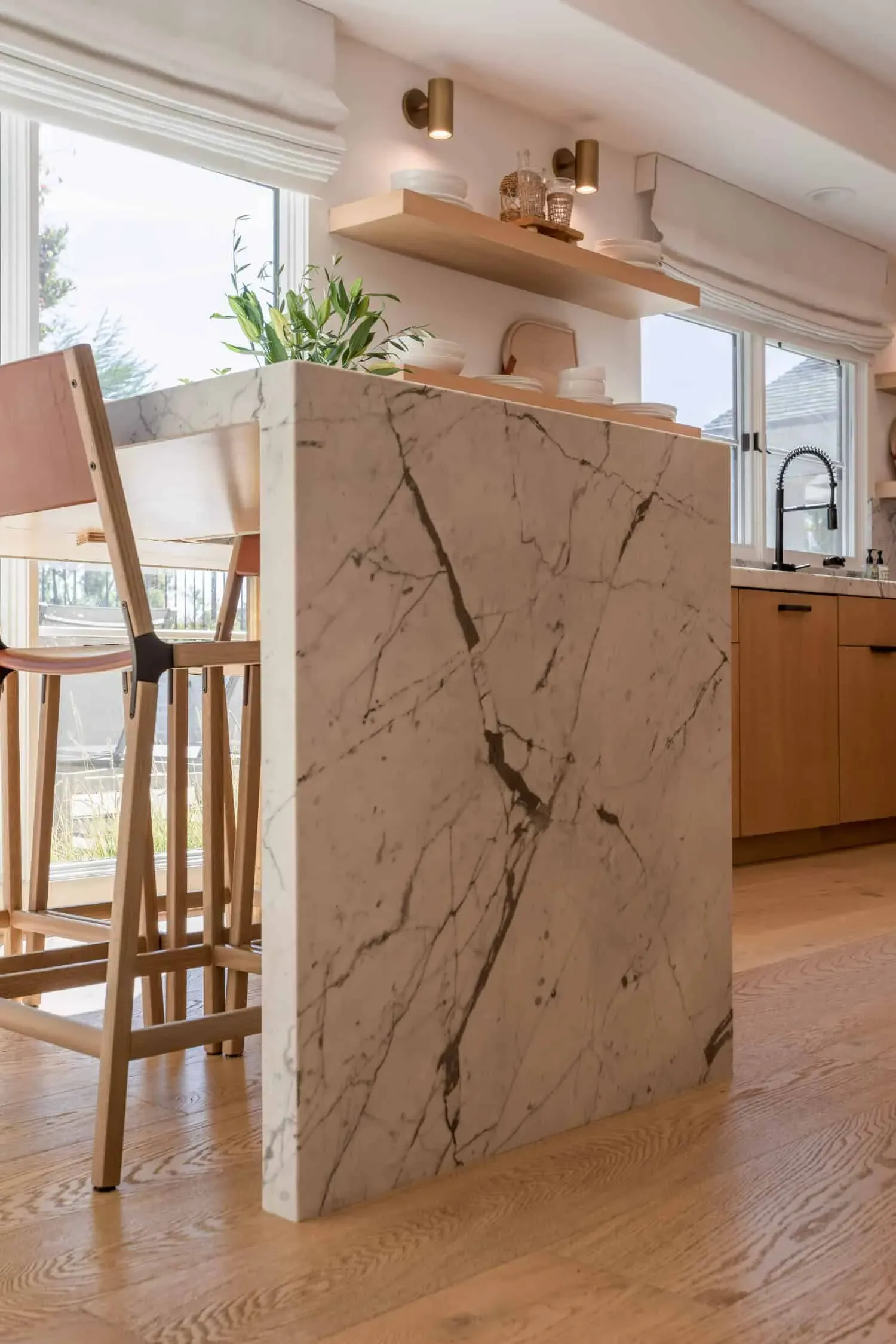 Veined marble waterfall island end meeting light oak cabinetry beside wooden bar stools
