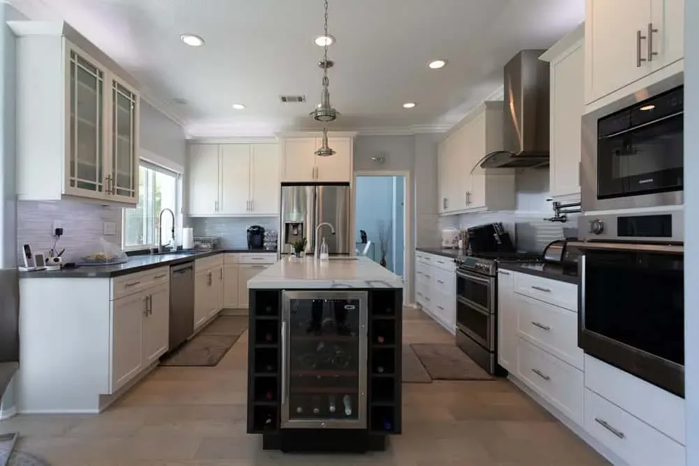 White shaker kitchen with a dark island housing a wine cooler, a stainless range and a marble-look backsplash