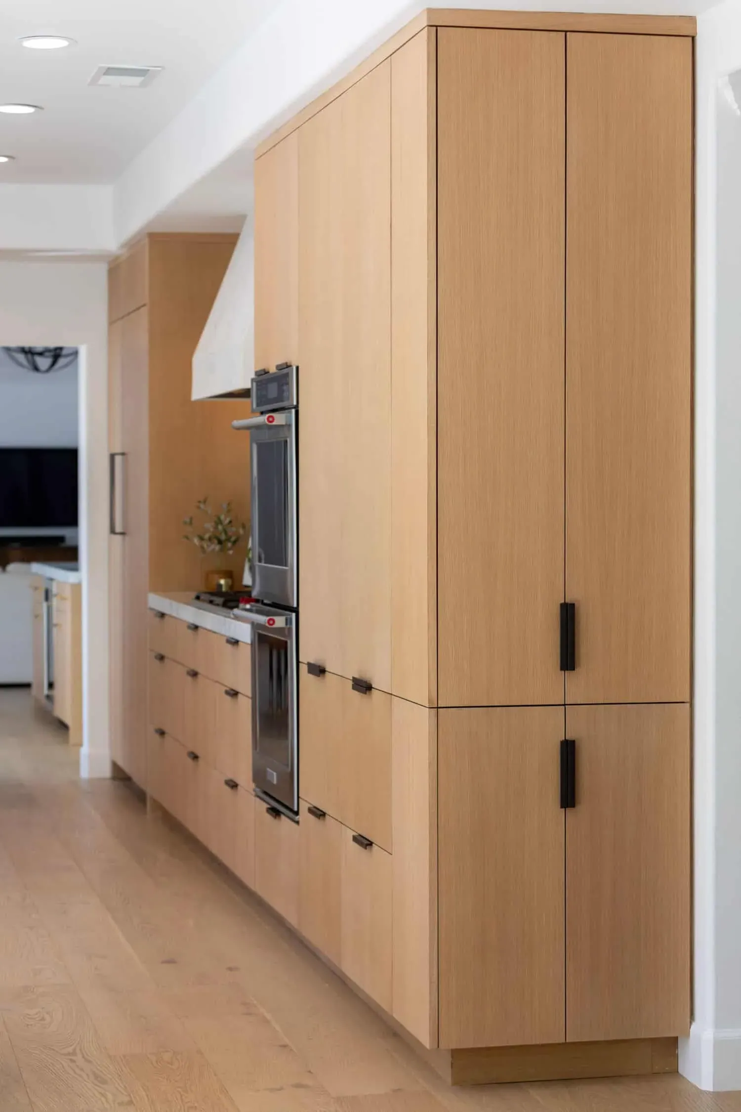 Flat oak cabinetry with integrated refrigerator panels and black pulls beside stacked wall ovens