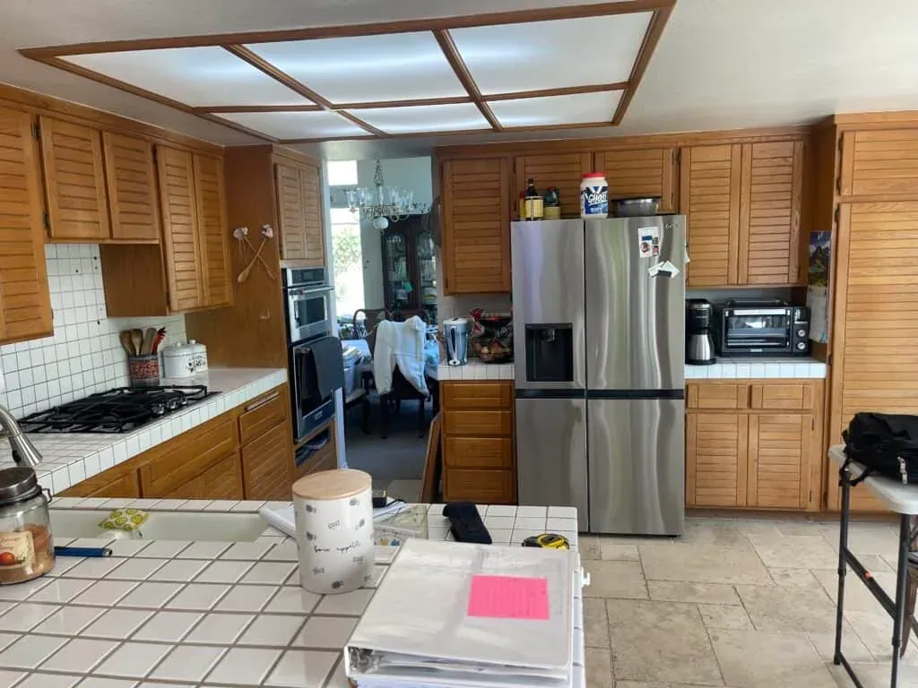 Original honey oak kitchen with louvered cabinet doors, white tile countertops and a framed fluorescent ceiling light