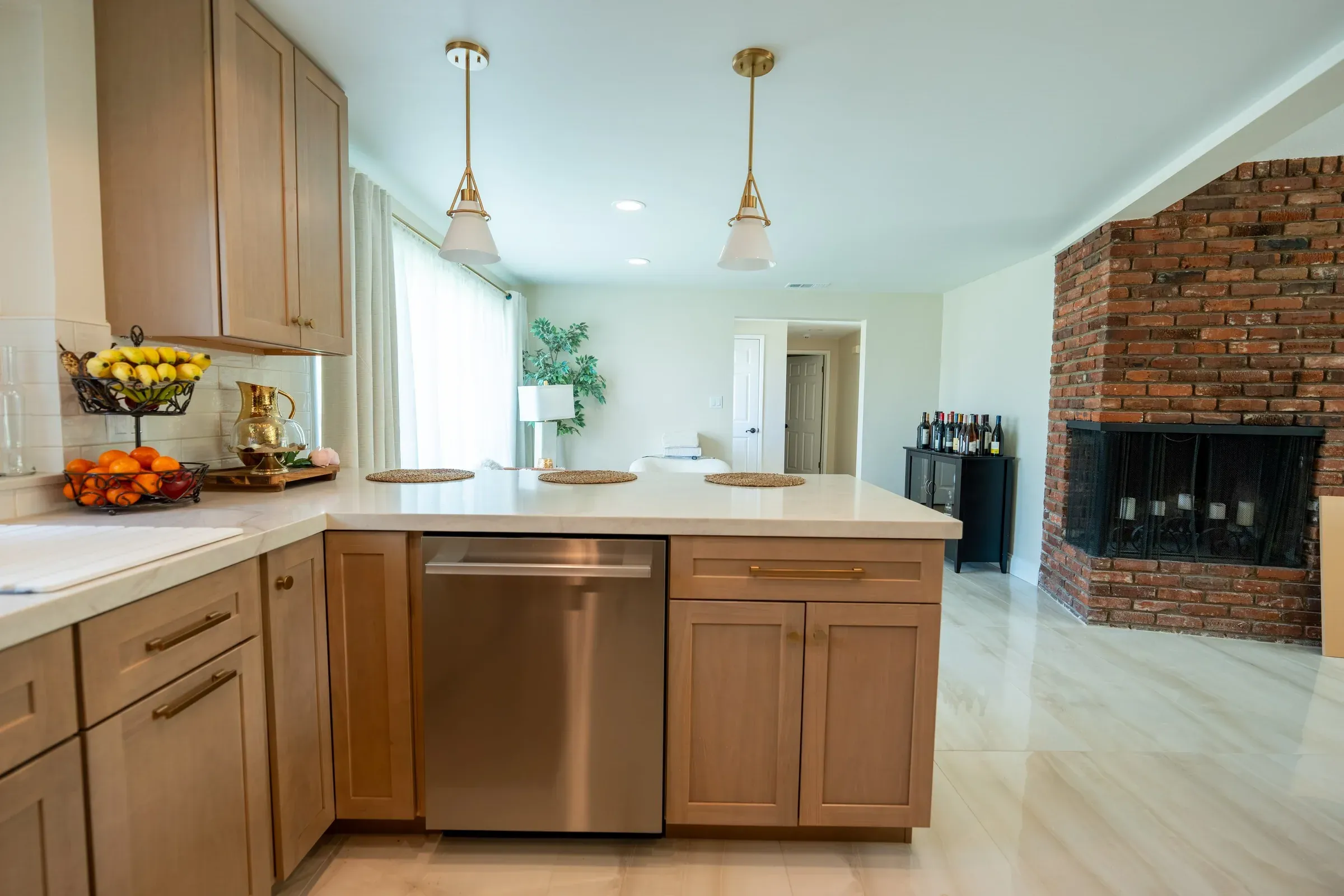 Oak shaker kitchen peninsula with a pale countertop, glass pendant lights, a stainless dishwasher and a brick fireplace beyond