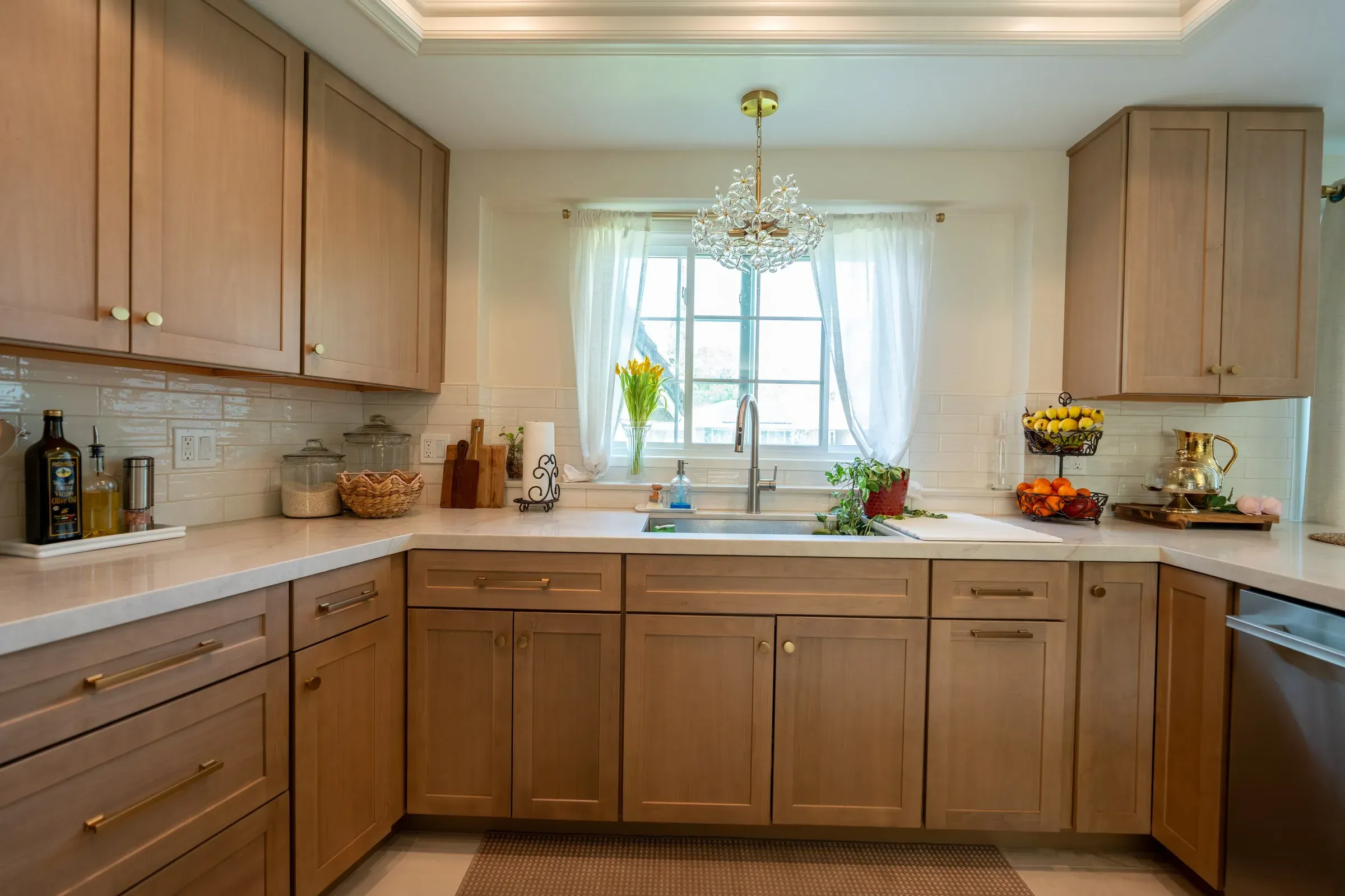 Light oak shaker cabinets with brass hardware around a sink under a window with a crystal chandelier