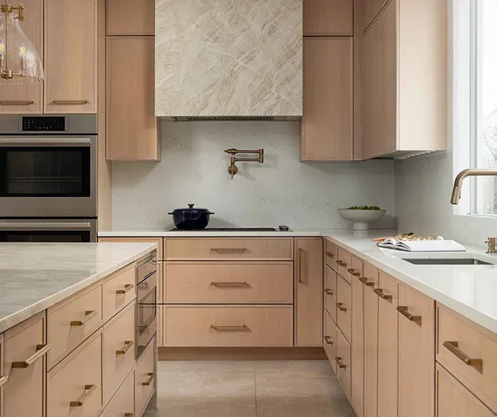 Omega Cabinetry photograph of a kitchen in its Jaxson door style in light oak with brass pulls and a stone hood, not a Kitchen Cabinets and Beyond project